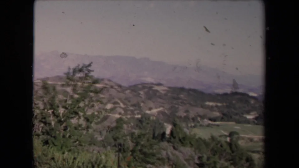 1967 Los Angeles-area hillside view with scrub vegetation, open green patches, distant ridges, and a pale hazy sky.