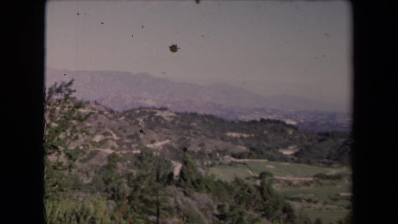 Hillside terrain with scrub, fields, and distant structures under hazy sky.