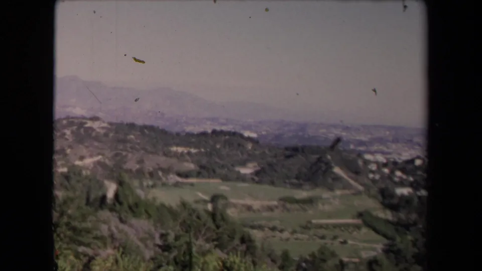 1967 Los Angeles-area panorama with a green valley floor, scrubby slopes, road lines, and distant haze over the hills.