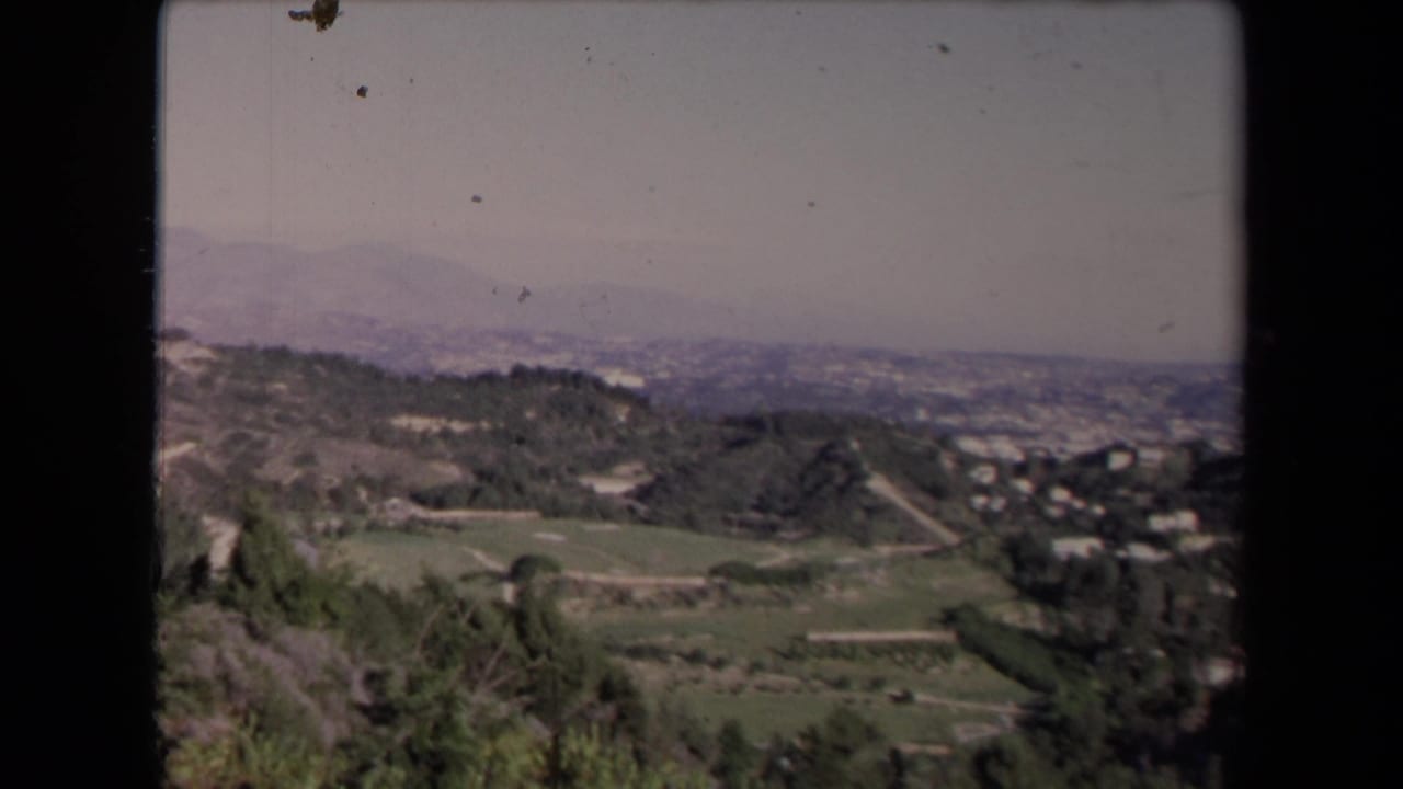 Wide hillside valley with green slopes, scattered homes, haze, and distant mountains.