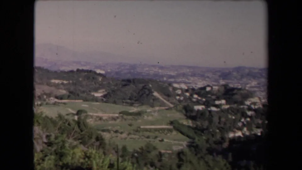 Los Angeles-area valley view from 1967 with winding paths, dark trees, hillside houses, and mountains under a pale sky.