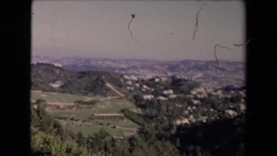 Mid-pan 8mm frame of a Los Angeles-area valley, with shrubs in front, hillside homes beyond, and hazy ridges on the horizon.