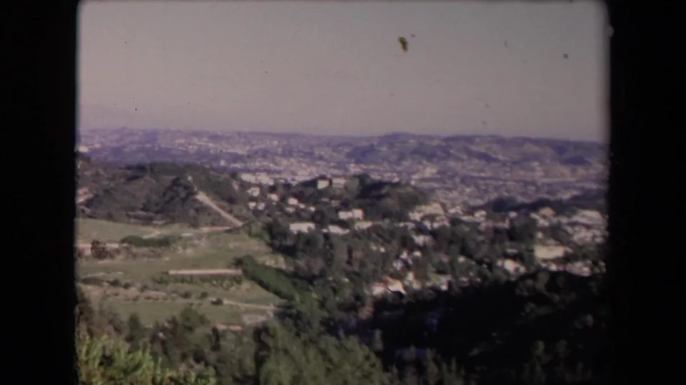 Final 1967 Los Angeles-area frame showing green hillside terrain, dark foreground trees, scattered residences, and hazy mountains.
