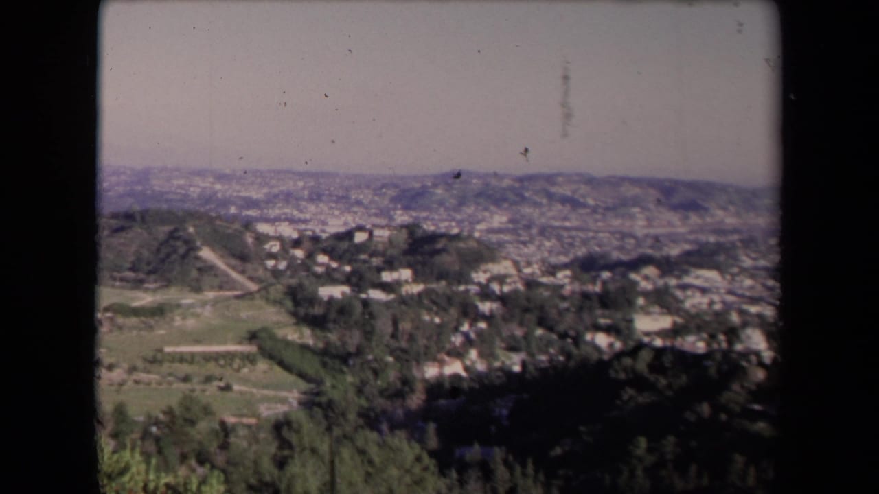 Elevated view over green hills, houses, distant city blocks, and hazy ridgelines.