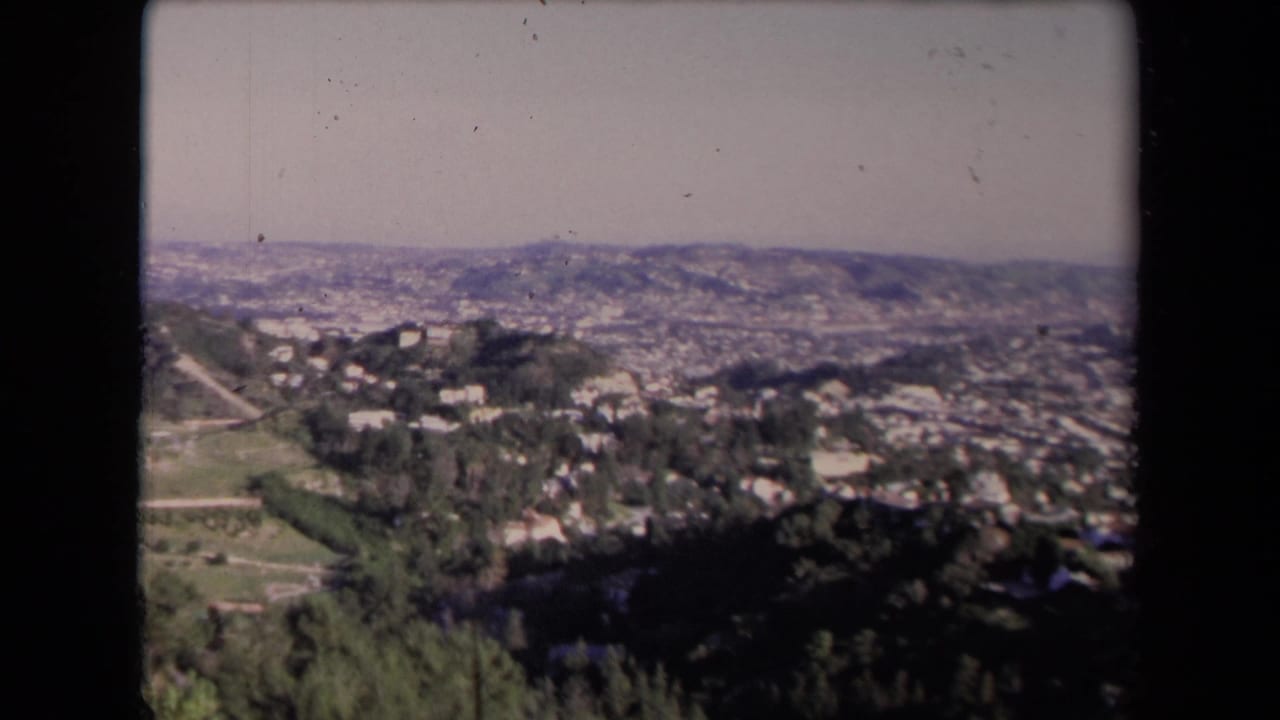 1967 Los Angeles frame with hillside homes and trees in the foreground and a broad, hazy city panorama beyond.
