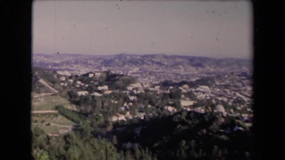 1967 Los Angeles frame with hillside homes and trees in the foreground and a broad, hazy city panorama beyond.