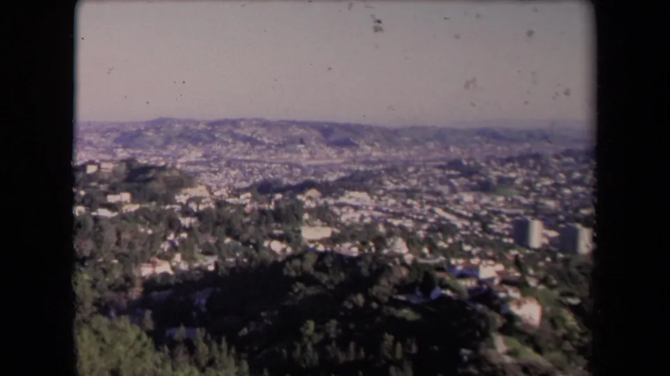 1967 Los Angeles frame with dark hillside foreground, dense residential areas, distant ridgelines, and a pale hazy sky.