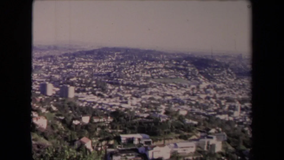 Elevated 8mm frame of Los Angeles hillside residences, greenery, low buildings, and a hazy skyline distance.