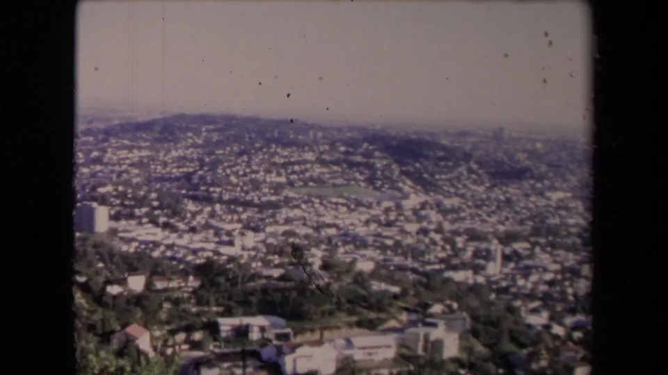 1967 home-movie frame showing Los Angeles neighborhoods below a high slope, with distant hills and soft film grain.