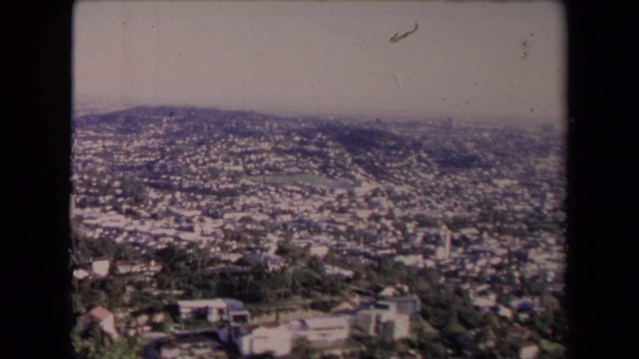 High-angle view over a sprawling city with hills, homes, trees, haze, and film-frame borders.