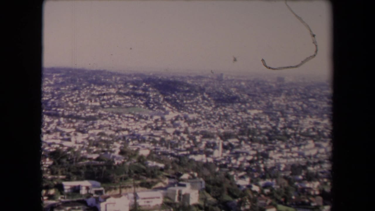 Wide city panorama with dense housing, hillside foreground, haze, dust, scratches, and dark film border.
