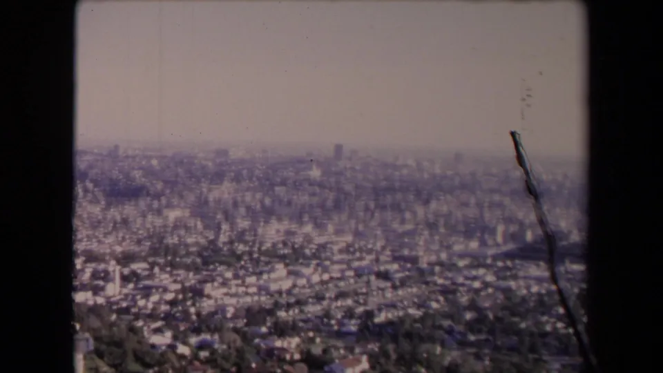 1967 8mm city overlook showing Los Angeles through haze, with foreground vegetation or a dark edge object crossing the frame.