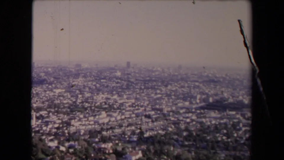 1967 Los Angeles city view from an overlook, showing dense neighborhoods under a pale hazy sky with 8mm film wear.
