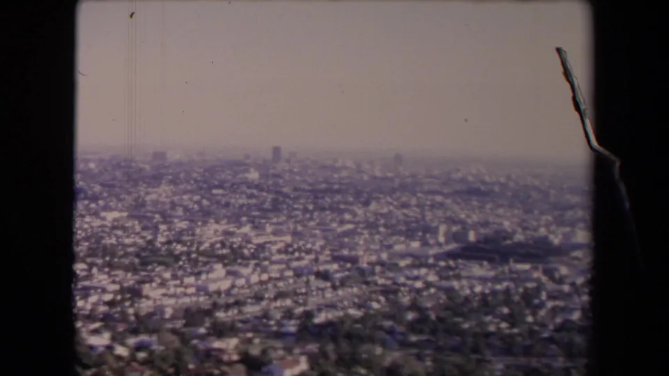 1967 Los Angeles home-movie frame showing a hazy elevated view across dense housing, trees, and distant high-rises.