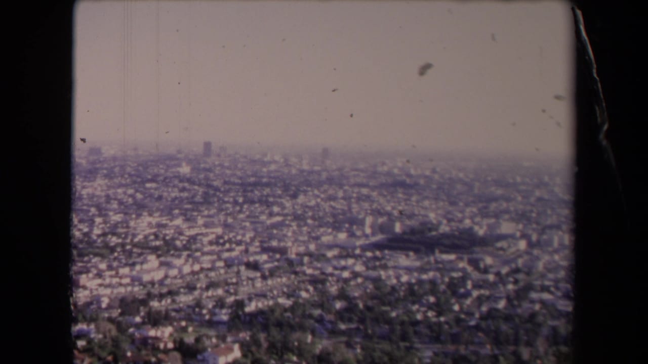 1967 Los Angeles 8mm frame with a wide hazy panorama, distant skyline forms, and dense low-rise urban blocks.