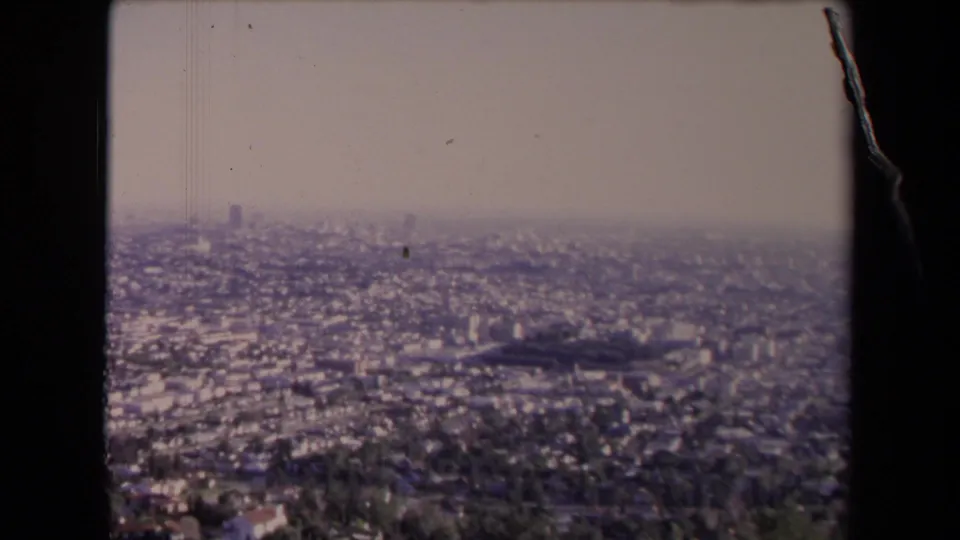 1967 Los Angeles frame looking over trees and dense city blocks toward a soft, hazy skyline.