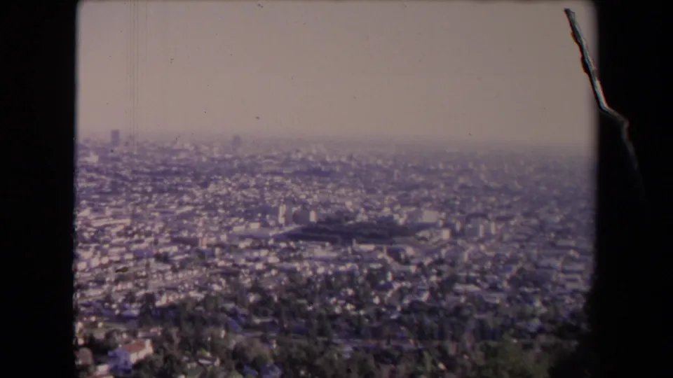 1967 Los Angeles home-movie frame showing a hazy panoramic view of dense buildings, tree cover, and distant skyline shapes.