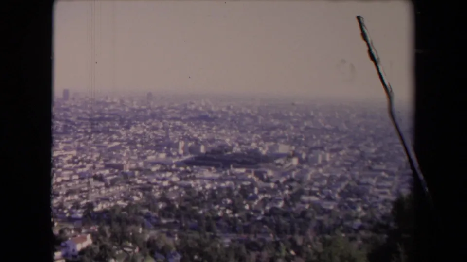 1967 8mm view from an elevated Los Angeles overlook, showing dense urban sprawl under a hazy sky with film border and edge scratch visible.