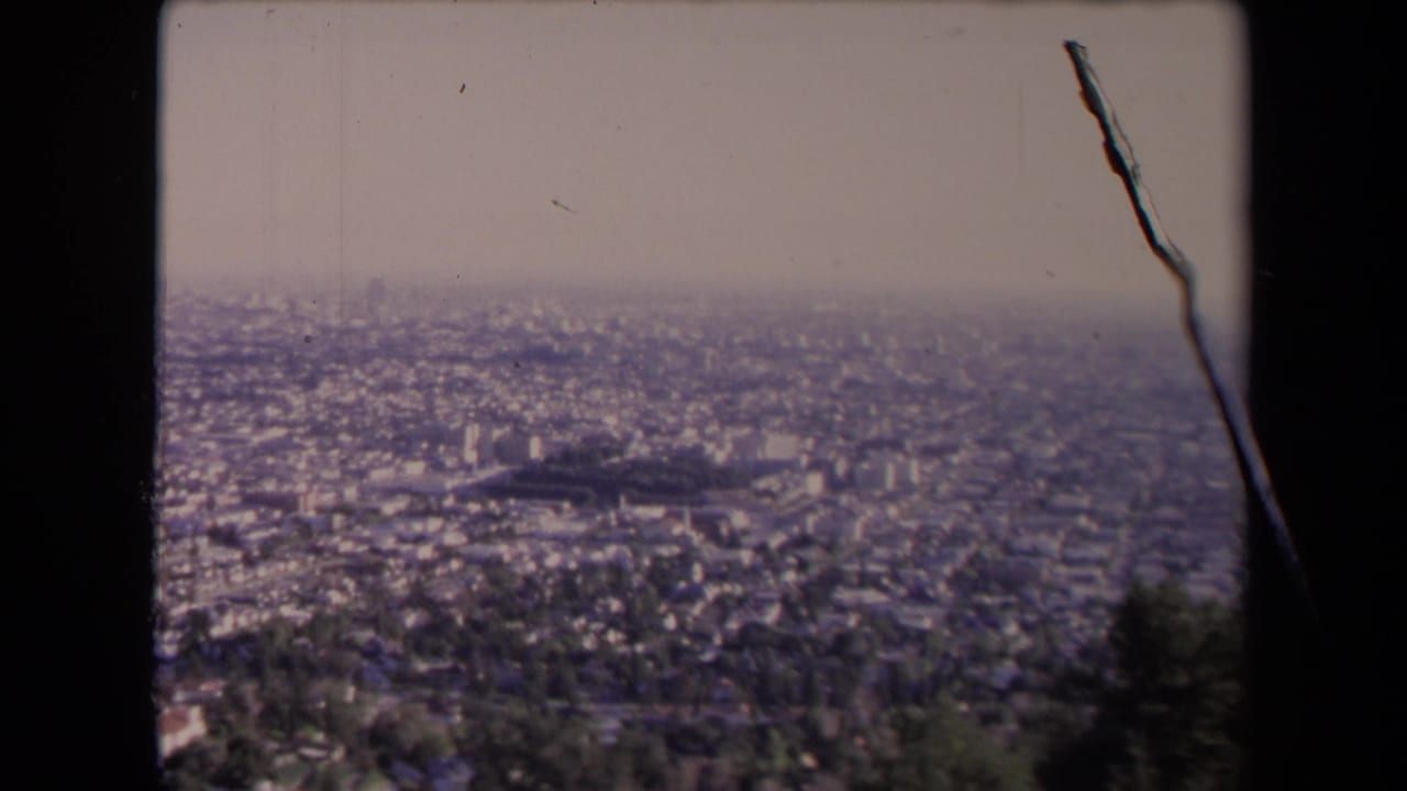 Elevated 1967 Los Angeles city view with low-rise neighborhoods, scattered taller buildings, haze, and visible 8mm film wear along the frame edge.