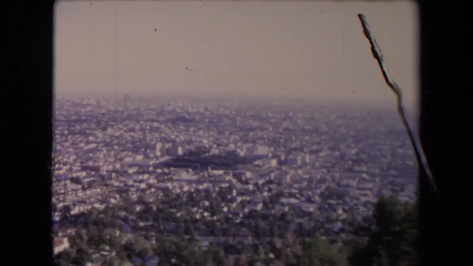 Elevated 1967 Los Angeles city view with low-rise neighborhoods, scattered taller buildings, haze, and visible 8mm film wear along the frame edge.
