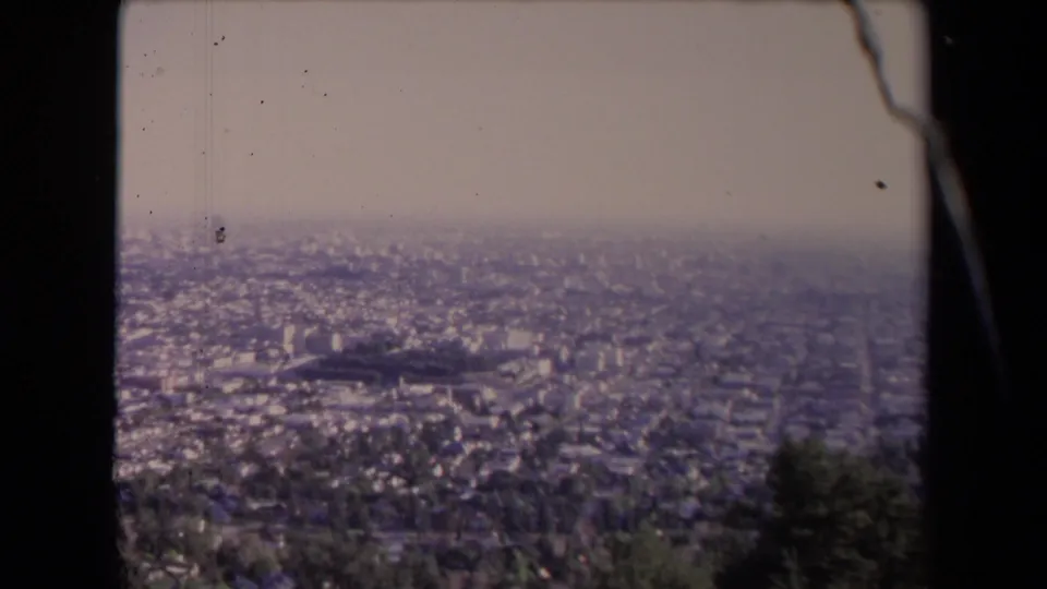 Late-frame 1967 8mm panorama over Los Angeles, showing dense city blocks beneath a pale sky with foreground foliage and visible film scratches.