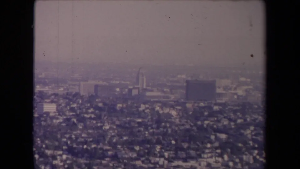 Muted 8mm frame of 1967 Los Angeles with a hazy skyline, broad residential area, and dark sprocket-side border visible.
