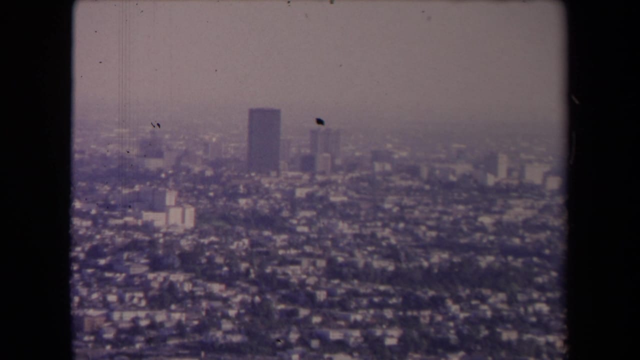 Hazy aerial view over dense Los Angeles neighborhoods with a dark high-rise tower in the distance.