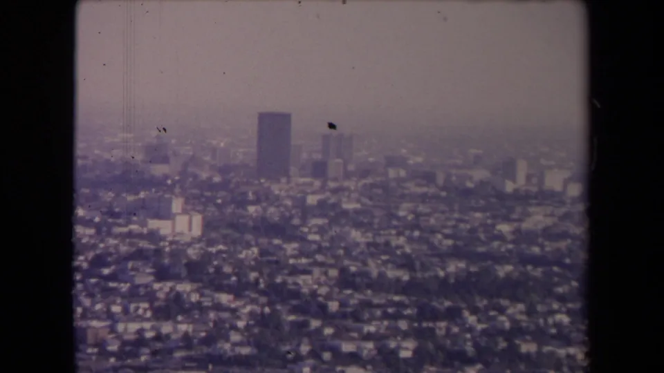 1967 Los Angeles aerial view showing dense residential blocks, distant high-rise buildings, haze, and visible 8mm film borders.