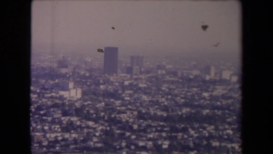 1967 Los Angeles skyline from above, with low-rise neighborhoods, several taller buildings, muted color, and 8mm film wear.