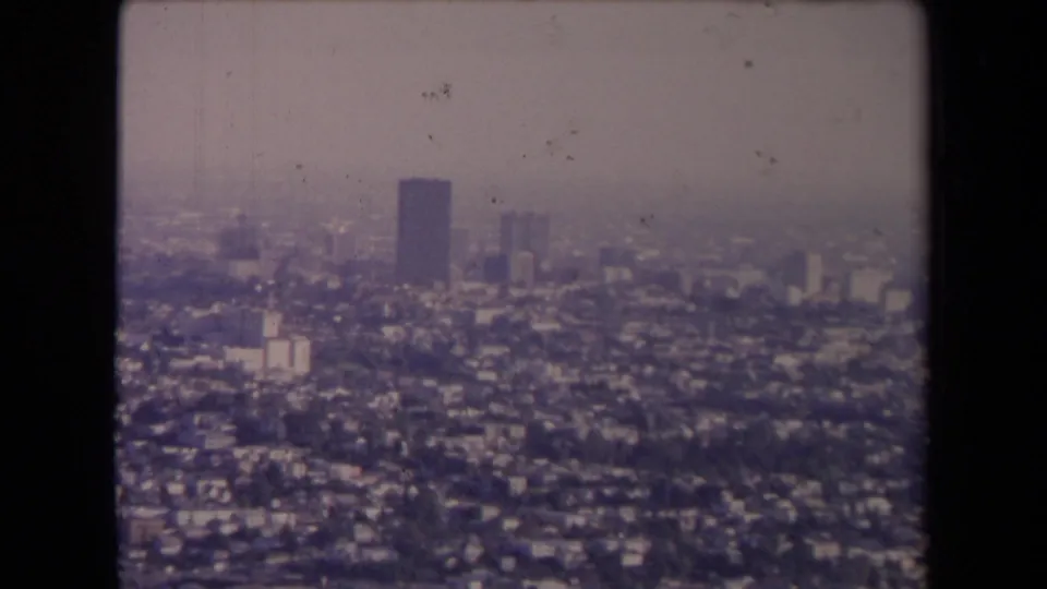 Mid-1960s Los Angeles aerial frame showing a sprawling urban field, distant towers, haze, and soft 8mm home-movie texture.