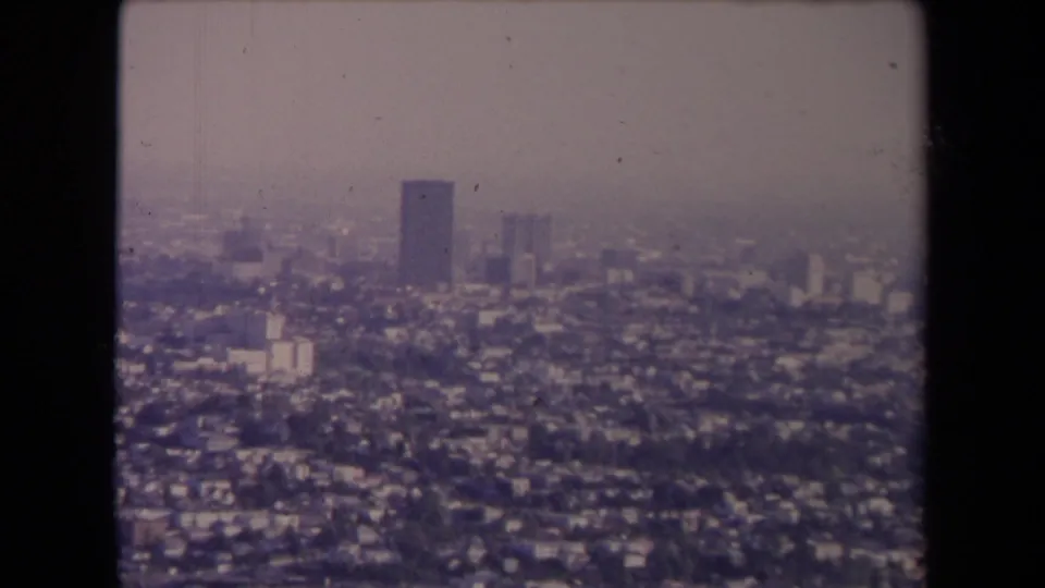1967 Los Angeles aerial cityscape with residential density, distant high-rises, hazy atmosphere, and visible 8mm frame boundaries.