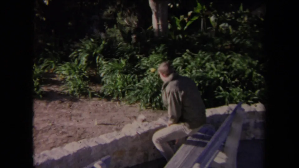 In 1967 Los Angeles home-movie footage, a man sits near a low stone wall with dense garden foliage behind him.