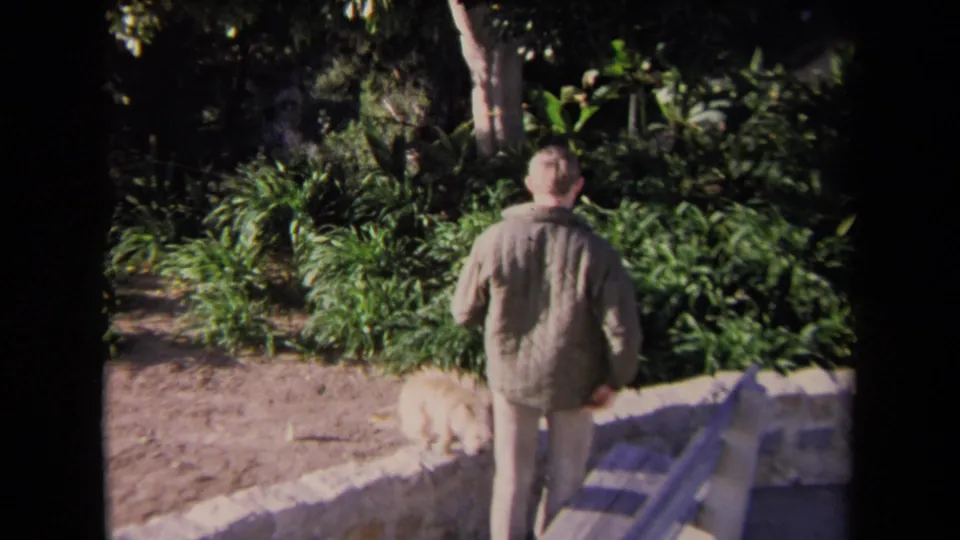 The man walks beside the low stone wall in a Los Angeles garden while a small light-colored dog moves near the plants.