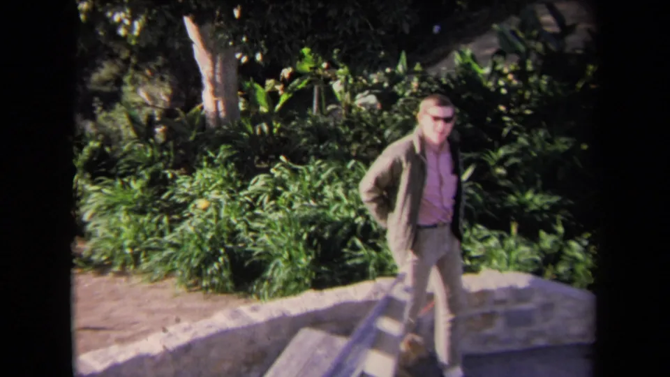 The man turns toward the camera in a 1967 Los Angeles garden scene, with the dog partly visible near the low wall.