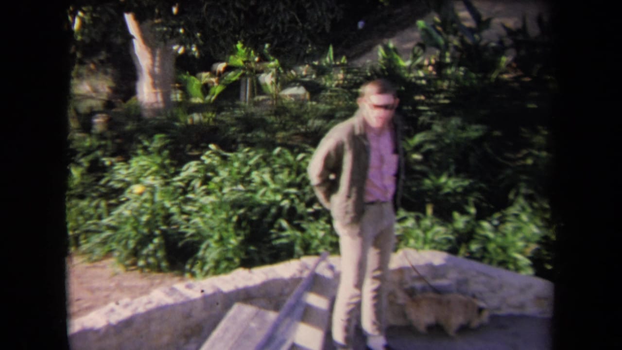 1967 Los Angeles home-movie frame showing a man in a pink shirt and jacket near stone steps and lush landscaping.