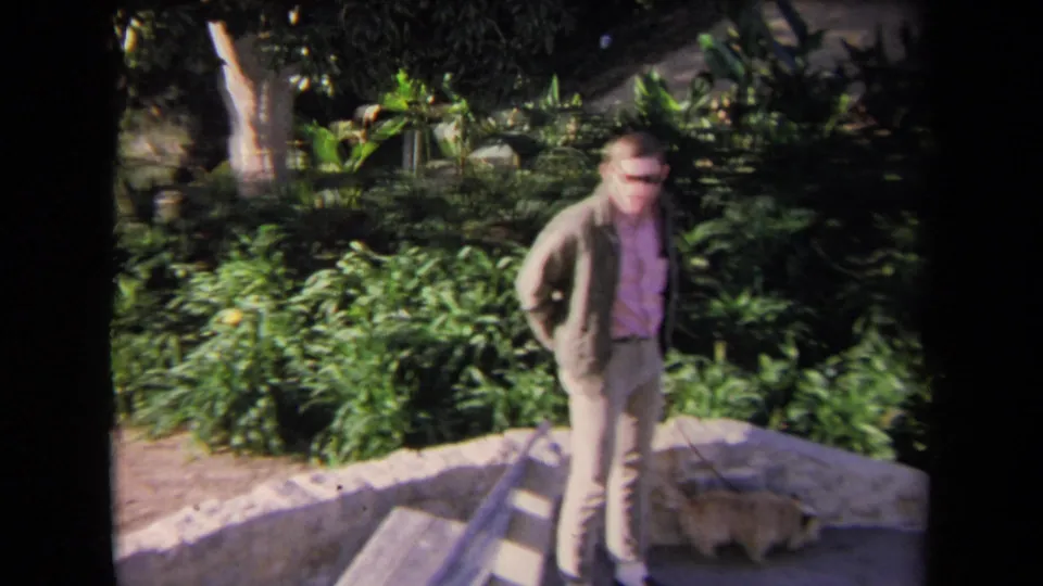 1967 Los Angeles home-movie frame showing a man in a pink shirt and jacket near stone steps and lush landscaping.