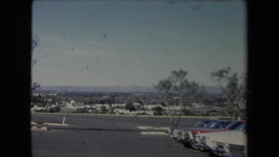 1967 Los Angeles view from a parking area toward houses, trees, parked cars, and a distant hazy horizon.