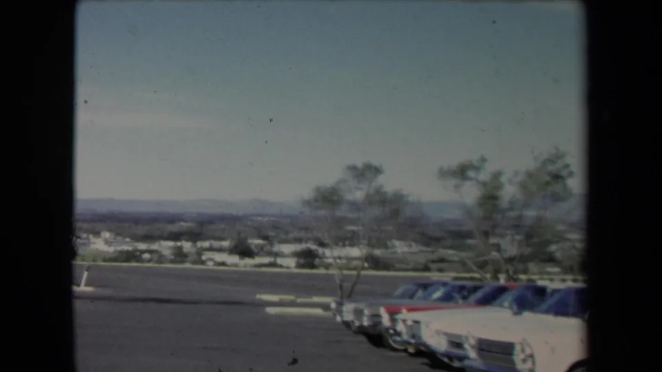 1967 Los Angeles-area 8mm frame showing parked mid-century cars along an overlook lot, with trees, suburban sprawl, and hills in the distance.