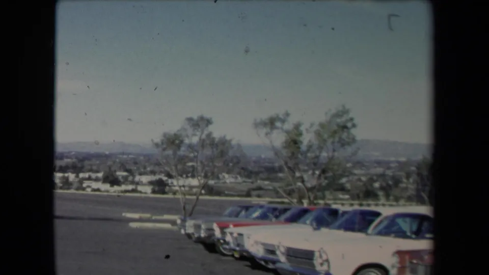 1967 Los Angeles-area frame with a row of parked cars at right, wind-shaped trees, a low suburban horizon, and hazy hills beyond.