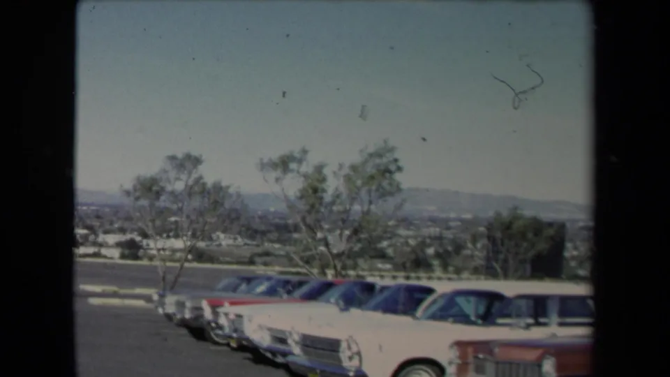 1967 8mm home-movie frame from Los Angeles showing mid-century cars in a parking area, sparse trees, and a broad distant hillside view.