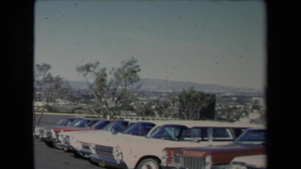 1967 Los Angeles-area frame with several parked sedans in the foreground, trees moving in the breeze, distant suburbs, and hazy hills.
