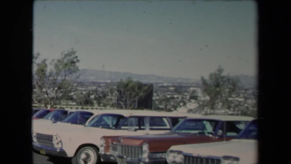 1967 Los Angeles home-movie frame with mid-century American cars parked in a row near a hillside overlook.