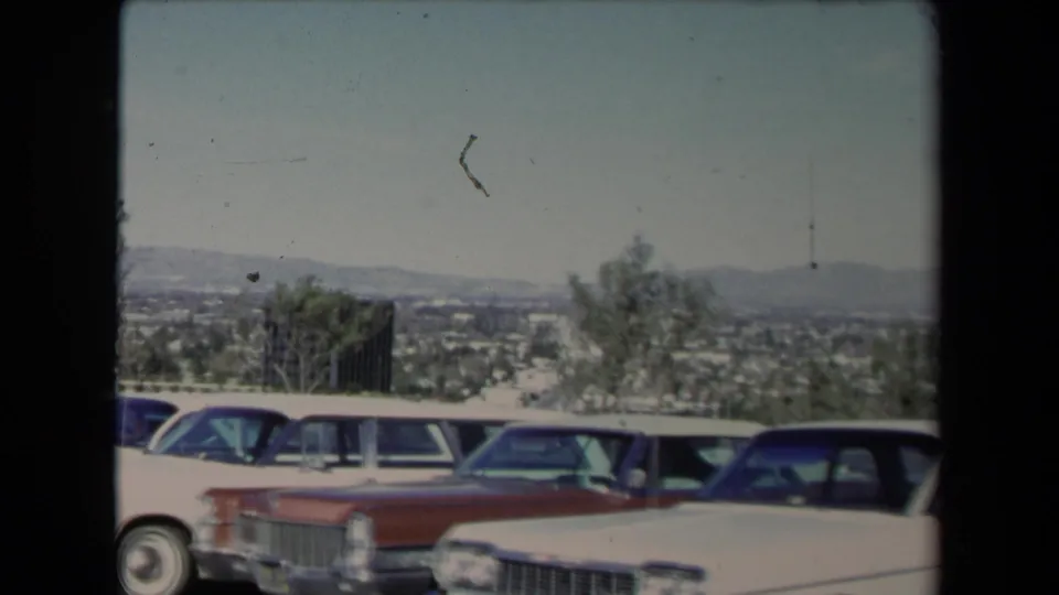 Soft 8mm frame of a Los Angeles hillside car lot with 1960s sedans, station wagons, trees, and distant hills.