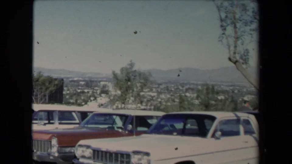 1967 Los Angeles 8mm frame showing classic cars at the edge of a hillside lot, with trees and the urban valley beyond.