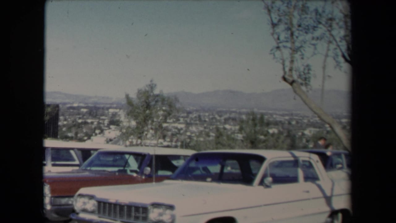 White sedan and red car parked at a Los Angeles overlook with city and hills beyond.