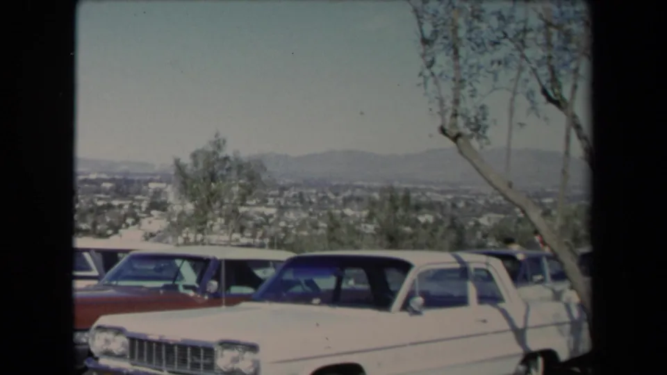 1967 Los Angeles overlook view with a white sedan, red vehicle, trees, and a hazy city basin in the background.