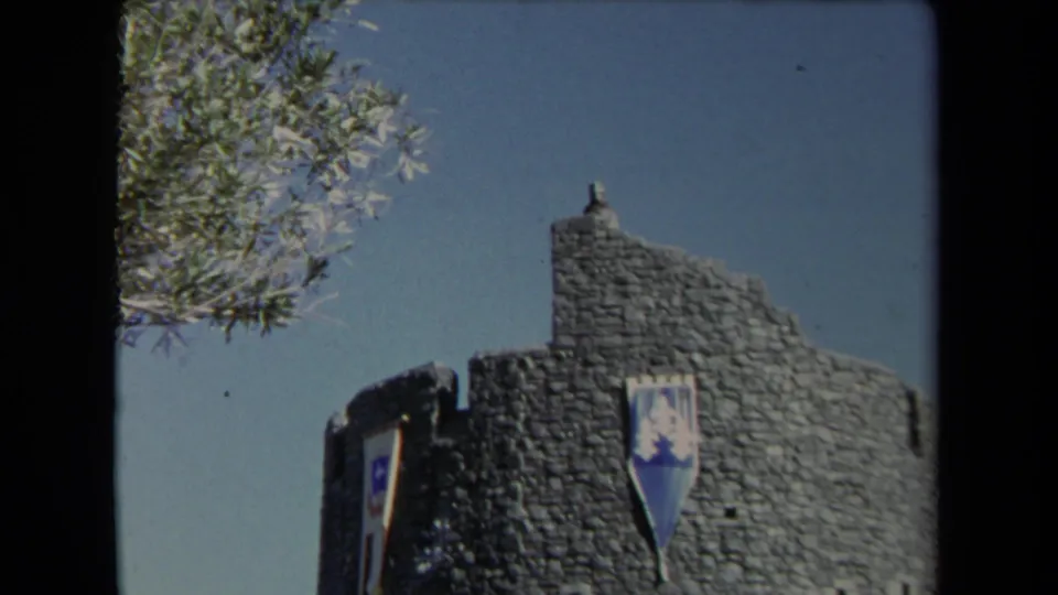 1967 Los Angeles 8mm frame focused on the Universal City Studios stone tower with a shield banner and tree branches nearby.