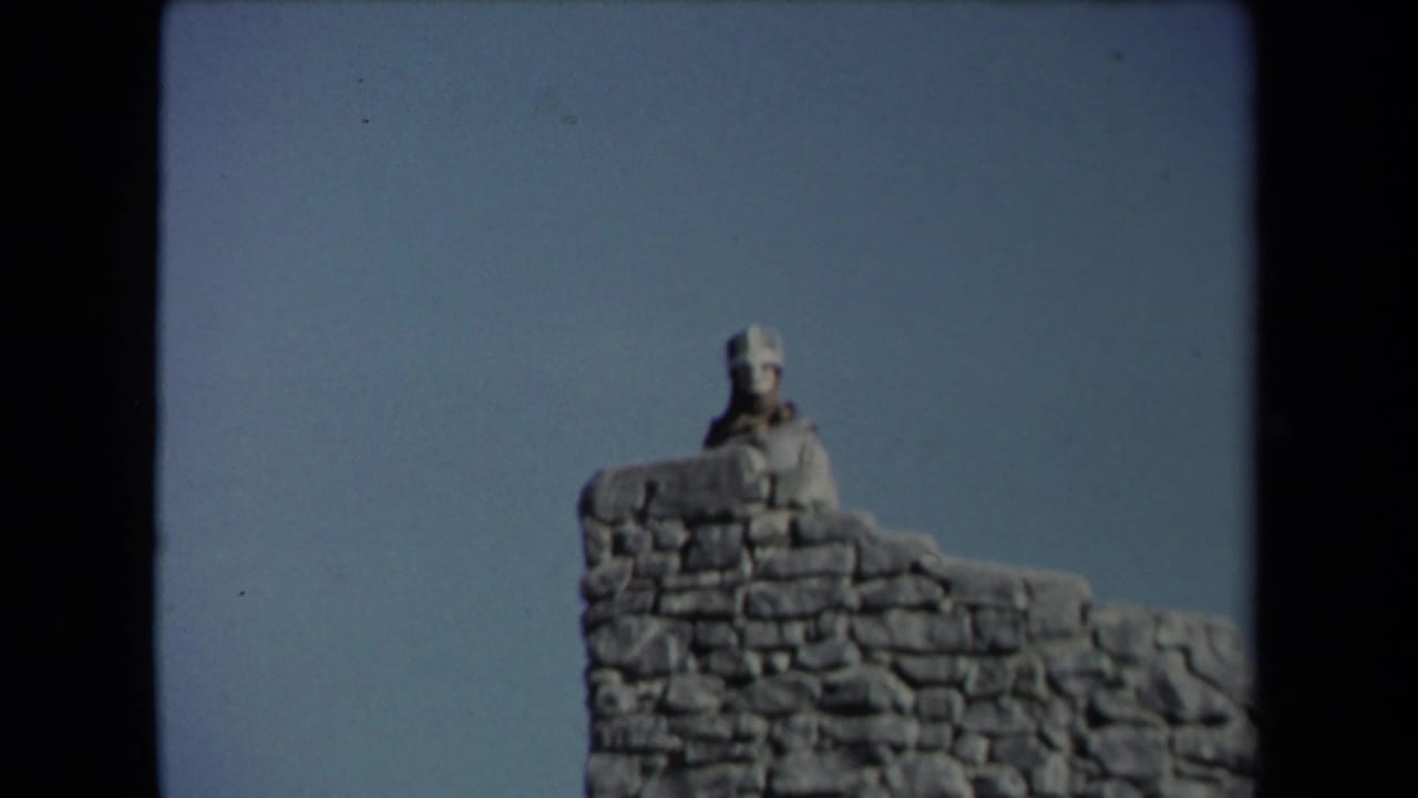 Pale statue or figure above a rough stone wall under a blue sky.