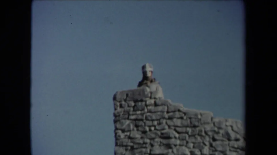 1967 Los Angeles 8mm frame showing a pale statue or figure rising behind a rough stone wall against blue sky.