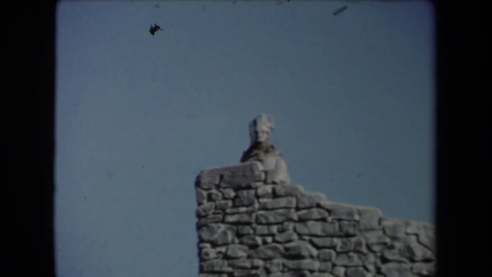 Outdoor 8mm frame from the supplied Los Angeles clip showing a pale figure behind a stone wall, with soft focus and blue sky.